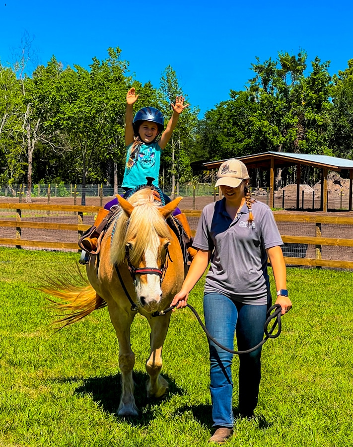 kid with hands up riding a horse being led by a trainer at Hidden Palms Ranch in Sanford, Florida
