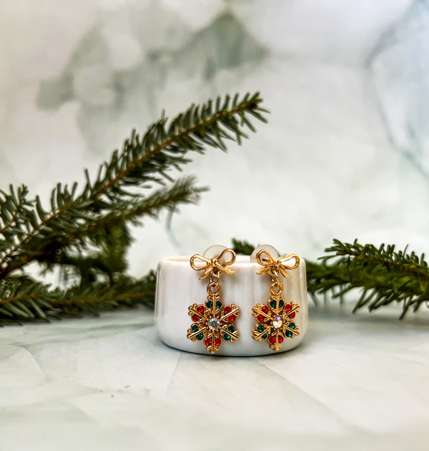 pair of Christmas earrings - snowflakes with red and green gemstones and one CZ in the center - the part that goes into the ear is a bow - they are leaning on a jewelry box with Christmas tree trimmings in the background