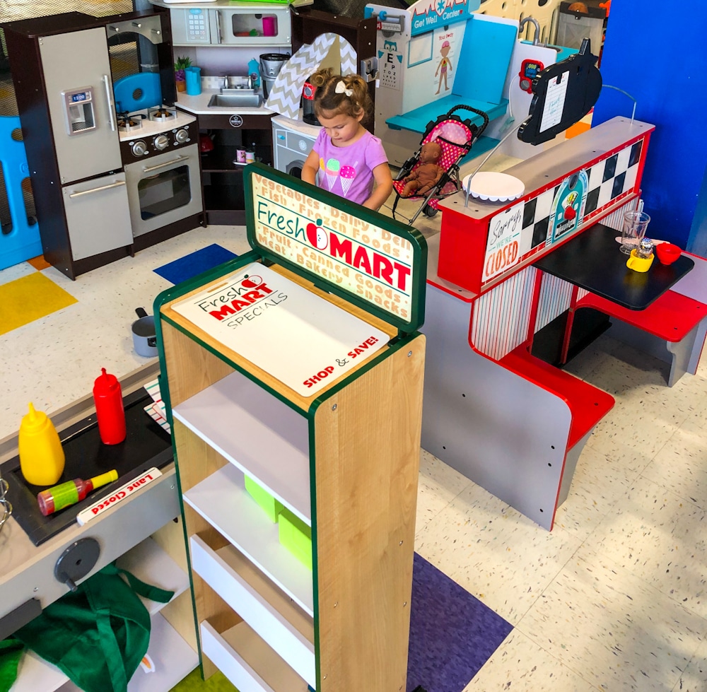 little girl playing in a pretend play area with kitchen, dinner, and grocery store sets